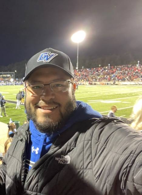 Hector Belmont smiling in Lubbers Stadium at a nighttime football game, sporting a GV hat